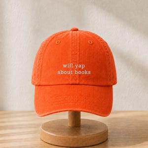 May include: A bright orange baseball cap featuring the white embroidered text will yap about books centered on the front panel. The cap has a structured crown, a curved brim, and a button at the top, showcasing a classic six-panel design.