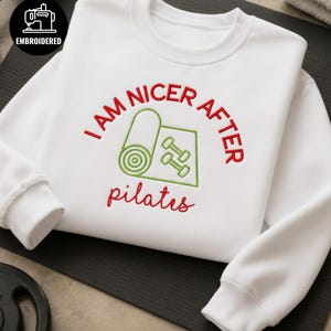 Puede incluir: Una sudadera blanca de manga larga con texto bordado en rojo que dice I AM NICER AFTER y pilates en letra cursiva. Entre el texto hay un gráfico bordado en verde de una esterilla de yoga enrollada y dos mancuernas pequeñas.