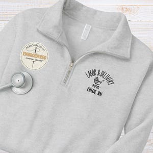 Könnte beinhalten: Ein hellgraues Sweatshirt mit Viertelreißverschluss, Kragen und Reißverschluss. Auf dem Sweatshirt steht der Text "LABOR & DELIVERY" mit einer Babywagen-Grafik und "CHLOE, RN" auf der Vorderseite. Ein kreisförmiger Aufnäher mit der Aufschrift "PERSONALIZE IT! EMBROIDERED COMFORT COLORS" befindet sich auf der linken Seite.