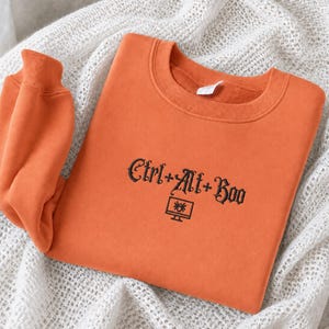 May include: An orange crewneck sweatshirt featuring black embroidered text that reads Ctrl+Alt+Boo, accompanied by a small icon of a computer monitor displaying a spider. The garment has long sleeves and a ribbed collar, cuffs, and hem.