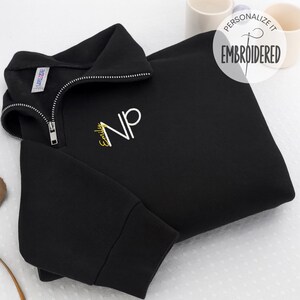 May include: Black quarter-zip sweatshirt with a silver zipper and embroidered white "NP" initials. The name "Emily" is embroidered in gold below the initials. The sweatshirt has a tag that says "Larkspur". The top right corner of the image has a white circle with the words "Personalize it" and "Embroidered".