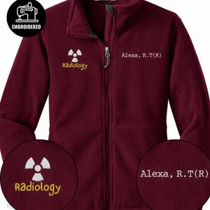 May include: A maroon fleece zip-up jacket featuring an embroidered white radiation symbol above the yellow text Radiology on the right chest and the white embroidered text Alexa, R.T(R) on the left chest, with a full-length front zipper.