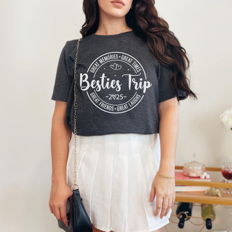 May include: Dark gray t-shirt with a white circular graphic that reads "Besties Trip -2025" surrounded by the phrases "Great Memories, Great Times, Great Friends, Great Laughs". The shirt is paired with a white pleated skirt.