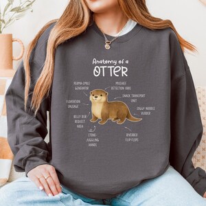 Puede incluir: Sudadera gris oscuro con una ilustraci&oacute;n de una nutria y etiquetas anat&oacute;micas como "Perma-Smile Generator" y "Riverbed Flip-Flops". El texto "Anatomy of an Otter" est&aacute; encima de la ilustraci&oacute;n. Un estilo casual y c&oacute;modo.