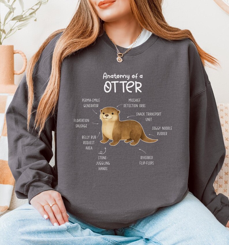 Puede incluir: Sudadera gris oscuro con una ilustraci&oacute;n de una nutria y etiquetas anat&oacute;micas como "Perma-Smile Generator" y "Riverbed Flip-Flops". El texto "Anatomy of an Otter" est&aacute; encima de la ilustraci&oacute;n. Un estilo casual y c&oacute;modo.