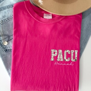 May include: A vibrant pink t-shirt with the word "PACU" in a floral design and the name "Hannah" in a script font. The shirt is folded and placed on top of a pair of blue jeans and a tan hat.