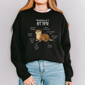 Puede incluir: Sudadera negra con un diagrama humor&iacute;stico de la anatom&iacute;a de una nutria. El dise&ntilde;o incluye etiquetas como "Perma-Smile Generator" y "Stone-Juggling Hands". La ilustraci&oacute;n de la nutria es en tonos marrones y beige.