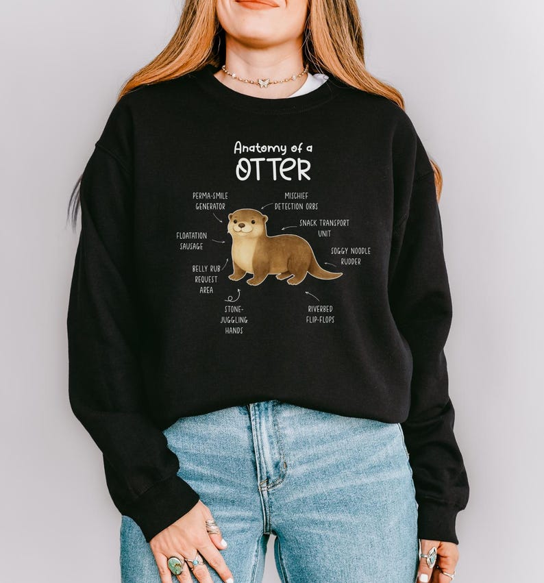 Puede incluir: Sudadera negra con un diagrama humor&iacute;stico de la anatom&iacute;a de una nutria. El dise&ntilde;o incluye etiquetas como "Perma-Smile Generator" y "Stone-Juggling Hands". La ilustraci&oacute;n de la nutria es en tonos marrones y beige.