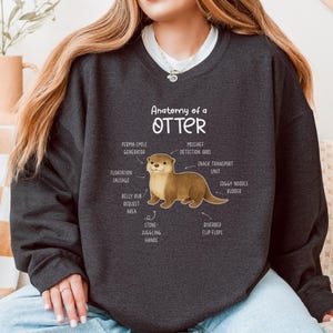 Puede incluir: Sudadera gris oscuro con una ilustraci&oacute;n de una nutria y etiquetas anat&oacute;micas como "Perma-Smile Generator" y "Riverbed Flip-Flops". El texto "Anatomy of a Otter" est&aacute; encima del gr&aacute;fico de la nutria. La sudadera es de cuello redondo.