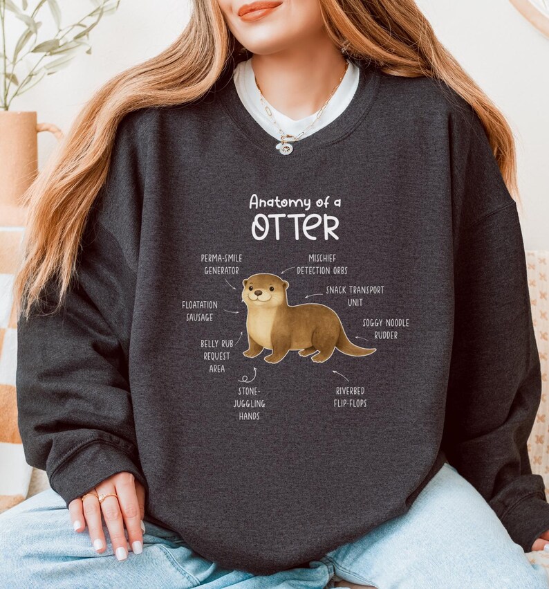 Puede incluir: Sudadera gris oscuro con una ilustraci&oacute;n de una nutria y etiquetas anat&oacute;micas como "Perma-Smile Generator" y "Riverbed Flip-Flops". El texto "Anatomy of a Otter" est&aacute; encima del gr&aacute;fico de la nutria. La sudadera es de cuello redondo.