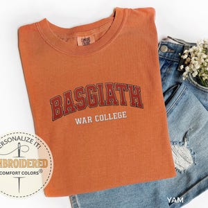 Puede incluir: Una camiseta naranja Comfort Colors con el texto bordado "BASGIATH WAR COLLEGE" en rojo y blanco. La camiseta est&aacute; doblada junto a unos pantalones cortos vaqueros azules con un peque&ntilde;o ramo de flores blancas.