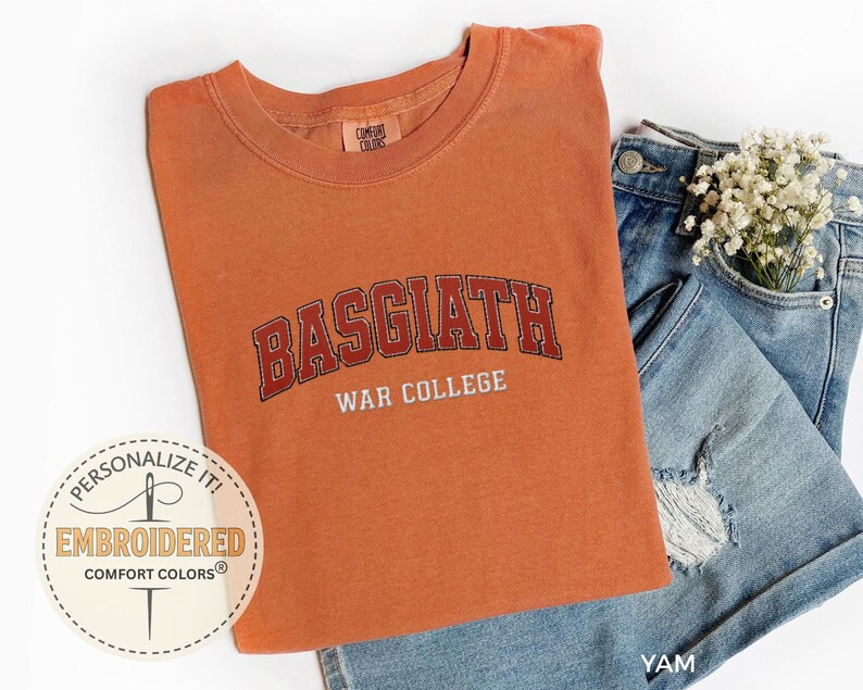 Puede incluir: Una camiseta naranja Comfort Colors con el texto bordado "BASGIATH WAR COLLEGE" en rojo y blanco. La camiseta est&aacute; doblada junto a unos pantalones cortos vaqueros azules con un peque&ntilde;o ramo de flores blancas.
