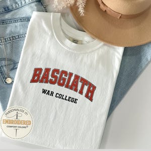 Puede incluir: Camiseta blanca con el texto "BASGIATH WAR COLLEGE" en letras rojas y negras. La camiseta est&aacute; doblada sobre unos vaqueros azules, con un sombrero color canela y un collar visible. Un logotipo circular con el texto "Personalize it! Embroidered Comfort Colors" est&aacute; en la esquina inferior izquierda.
