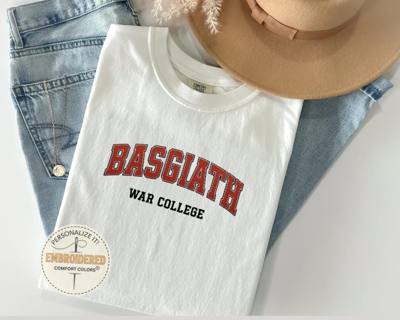 Puede incluir: Camiseta blanca con el texto "BASGIATH WAR COLLEGE" en letras rojas y negras. La camiseta est&aacute; doblada sobre unos vaqueros azules, con un sombrero color canela y un collar visible. Un logotipo circular con el texto "Personalize it! Embroidered Comfort Colors" est&aacute; en la esquina inferior izquierda.