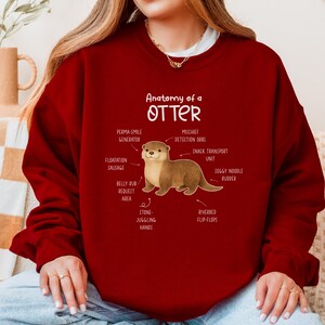 Puede incluir: Una sudadera burdeos con una ilustraci&oacute;n de una nutria y etiquetas anat&oacute;micas como "Perma-Smile Generator" y "Mischief Detection Orbs". El texto "Anatomy of a Otter" est&aacute; encima de la ilustraci&oacute;n.