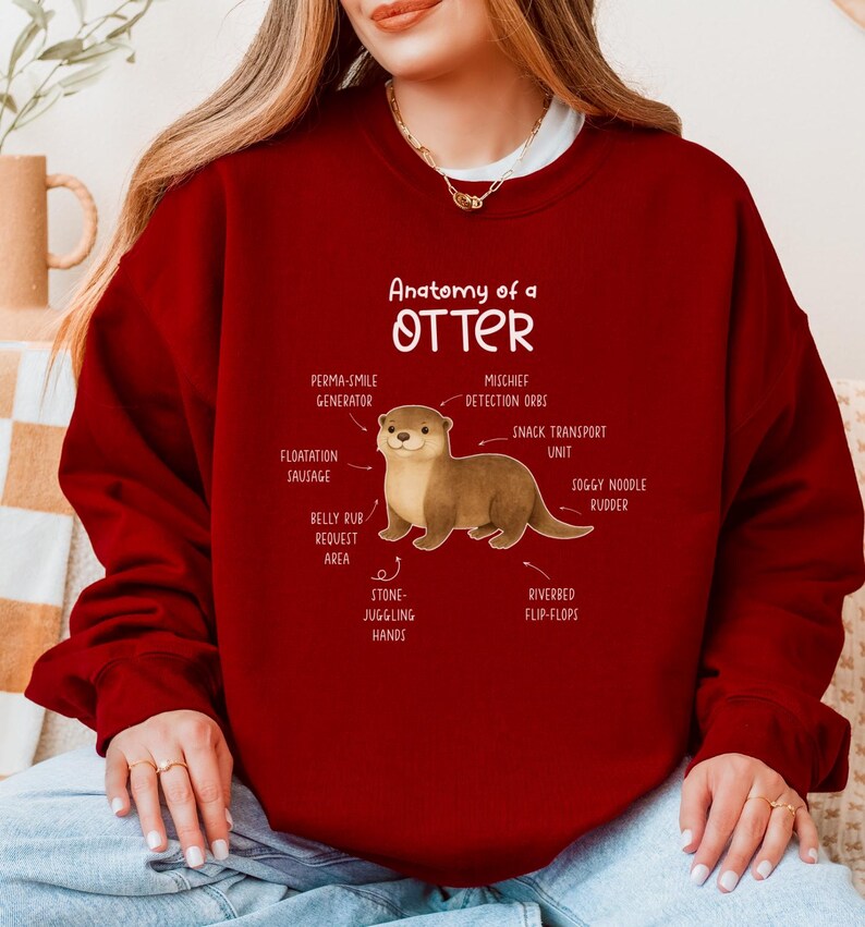 Puede incluir: Una sudadera burdeos con una ilustraci&oacute;n de una nutria y etiquetas anat&oacute;micas como "Perma-Smile Generator" y "Mischief Detection Orbs". El texto "Anatomy of a Otter" est&aacute; encima de la ilustraci&oacute;n.