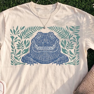 Blue linocut alligator head print surrounded by teal botanical foliage on white t-shirt, vintage reptile wildlife design
