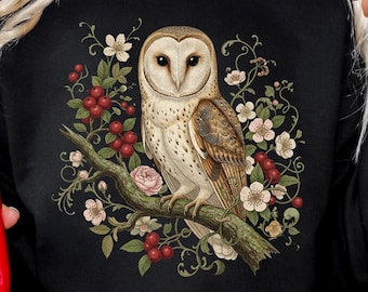 Puede incluir: Sudadera negra con una ilustración detallada de un búho posado en una rama, rodeado de flores y bayas. El búho tiene plumas marrones y blancas. El diseño está centrado en la parte delantera de la parte superior de manga larga.