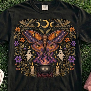 Peut inclure: Un t-shirt noir avec un motif central représentant un grand papillon orange et violet au-dessus d'un chaudron dégageant de la fumée violette. Le design inclut deux petits fantômes, des bougies allumées, des croissants de lune, des chauves-souris et des vignes florales.