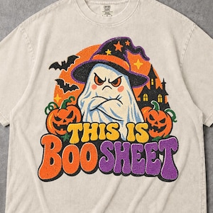 May include: A beige short-sleeve t-shirt featuring a graphic of a grumpy ghost wearing a witch hat, flanked by two jack-o'-lanterns, bats, and a haunted house silhouette. The text reads THIS IS BOOSHEET in orange and purple bubble letters.