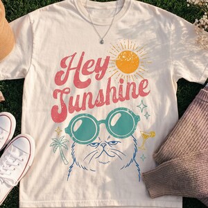 May include: Cream-colored t-shirt with the text "Hey Sunshine" and a graphic of a cat wearing sunglasses. The image also includes white sneakers, a brown corduroy hat, hair clips, and a mauve sweater.