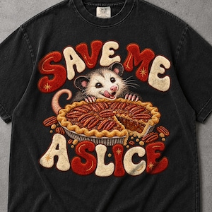 Puede incluir: Una camiseta negra de manga corta con un gráfico de una zarigüeya detrás de un pastel de nuez. El texto "SAVE ME A SLICE" está impreso en letras grandes y llamativas de color rojo y crema sobre y debajo de la zarigüeya. Incluye estrellas.