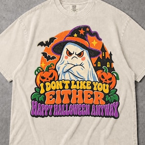 Könnte beinhalten: Ein beiges T-Shirt mit kurzen Ärmeln und einer Grafik eines mürrischen Geistes mit Hexenhut, umgeben von Kürbissen, Fledermäusen und einem Spukhaus. Der Text lautet: I DON'T LIKE YOU EITHER HAPPY HALLOWEEN ANYWAY in Gelb, Orange und Lila.