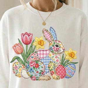 Può includere: Felpa bianca con ricamo di un coniglietto pasquale. Il design presenta un coniglio patchwork, tulipani, narcisi e uova di Pasqua decorate. Il coniglio è al centro del design.