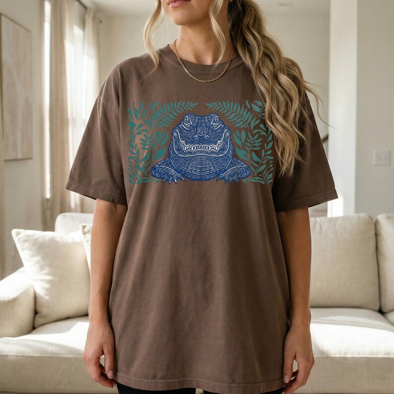 K&ouml;nnte beinhalten: Braunes T-Shirt mit einem blauen Alligator-Design, umgeben von gr&uuml;nen Bl&auml;ttern. Die Grafik befindet sich mittig auf der Vorderseite des Shirts. Das Shirt hat kurze &Auml;rmel und eine lockere Passform.