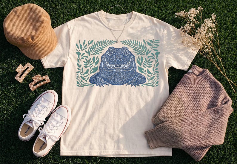 Blue linocut alligator head print surrounded by teal botanical foliage on white t-shirt, vintage reptile wildlife design