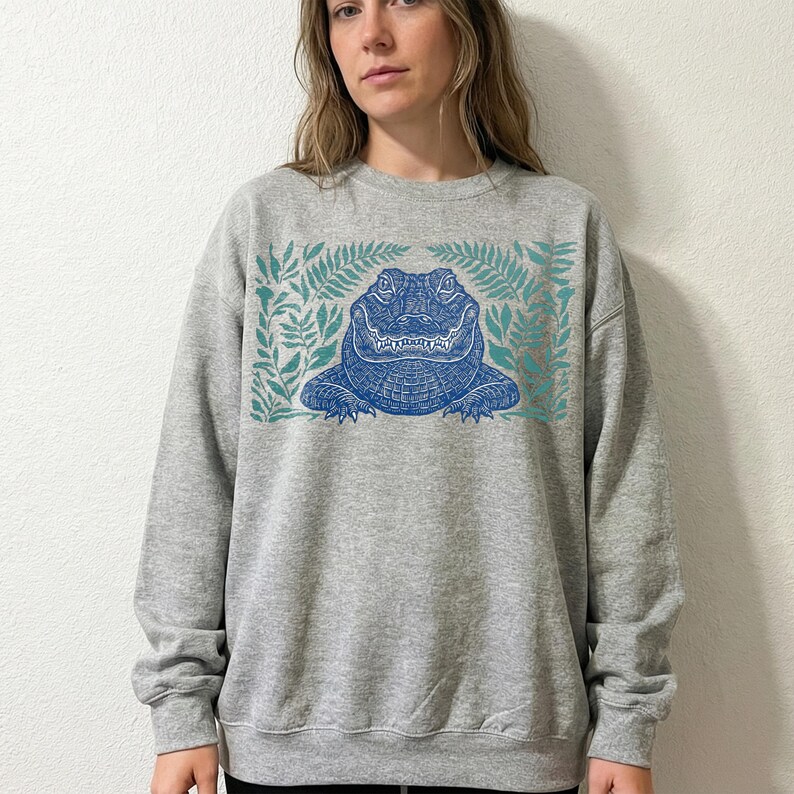 K&ouml;nnte beinhalten: Graues Sweatshirt mit einem blauen Alligator-Motiv, das von t&uuml;rkisfarbenen Farnbl&auml;ttern umrahmt wird. Der Alligator ist mittig platziert, mit detaillierten Schuppen und einem breiten Grinsen. Das Sweatshirt hat einen Rundhalsausschnitt und lange &Auml;rmel.