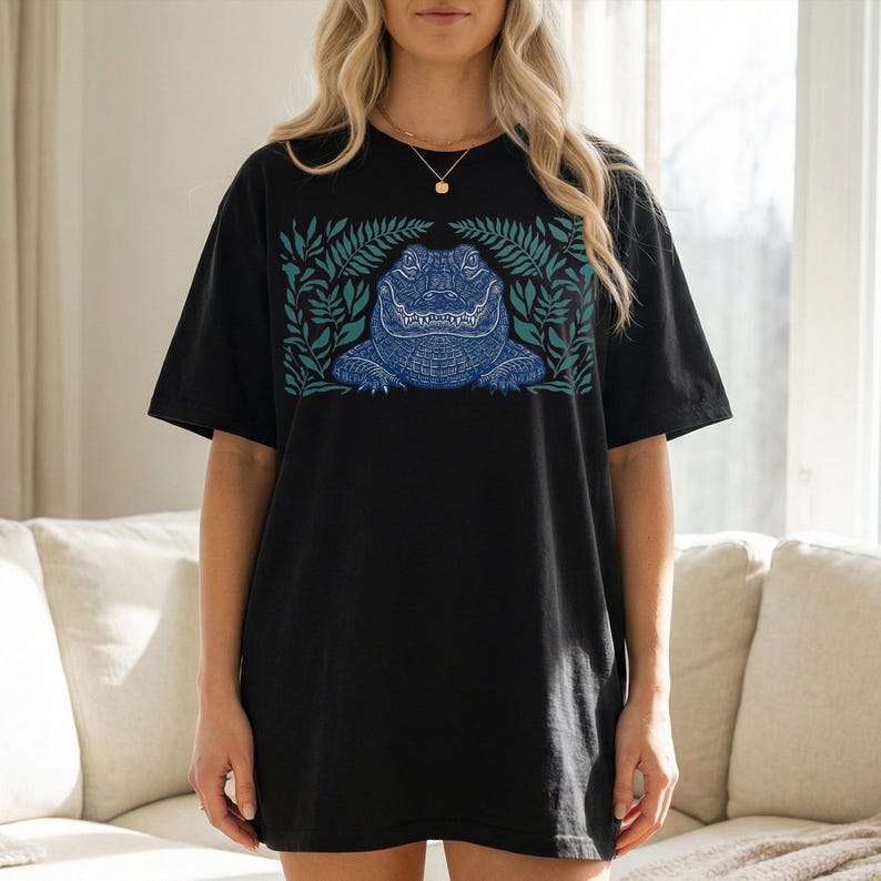 K&ouml;nnte beinhalten: Schwarzes T-Shirt mit einem blauen Alligator-Design, das von gr&uuml;nen Bl&auml;ttern umrahmt wird. Der Alligator ist mit komplizierten Mustern versehen. Das T-Shirt hat eine l&auml;ssige, &uuml;bergro&szlig;e Passform, die f&uuml;r den Alltag geeignet ist.