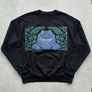 K&ouml;nnte beinhalten: Schwarzes Sweatshirt mit einem blauen Alligator-Design, umgeben von t&uuml;rkisfarbenen Bl&auml;ttern. Die Alligator-Illustration weist detaillierte Muster auf, und die Bl&auml;tter bilden einen Rahmen. Rundhals-Sweatshirt.