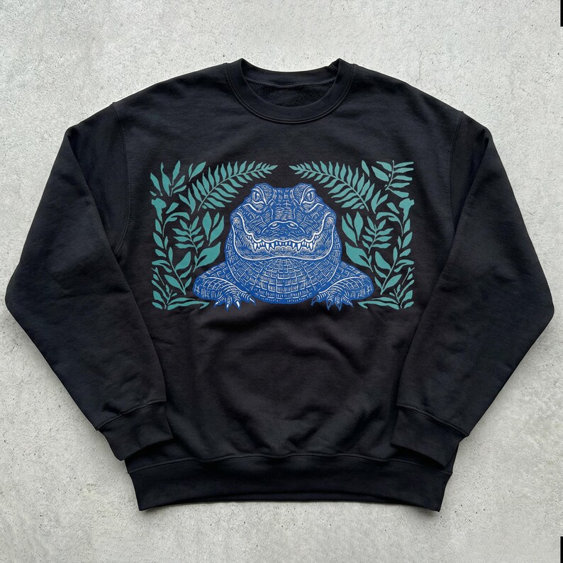 K&ouml;nnte beinhalten: Schwarzes Sweatshirt mit einem blauen Alligator-Design, umgeben von t&uuml;rkisfarbenen Bl&auml;ttern. Die Alligator-Illustration weist detaillierte Muster auf, und die Bl&auml;tter bilden einen Rahmen. Rundhals-Sweatshirt.