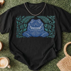 K&ouml;nnte beinhalten: Schwarzes T-Shirt mit einem blauen Alligatorendesign, umgeben von gr&uuml;nen Bl&auml;ttern. Der Alligator hat detaillierte Schuppen und einen strengen Ausdruck. Eine silberne Kette ist am Ausschnitt des Shirts drapiert. Das Shirt wird auf einer gr&uuml;nen Oberfl&auml;che mit Kaffeetassen und Accessoires pr&auml;sentiert.