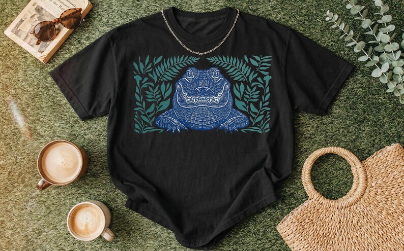 K&ouml;nnte beinhalten: Schwarzes T-Shirt mit einem blauen Alligatorendesign, umgeben von gr&uuml;nen Bl&auml;ttern. Der Alligator hat detaillierte Schuppen und einen strengen Ausdruck. Eine silberne Kette ist am Ausschnitt des Shirts drapiert. Das Shirt wird auf einer gr&uuml;nen Oberfl&auml;che mit Kaffeetassen und Accessoires pr&auml;sentiert.