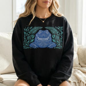 K&ouml;nnte beinhalten: Schwarzes Sweatshirt mit einem detaillierten blauen Alligator-Motiv, umgeben von t&uuml;rkisfarbenen Bl&auml;ttern. Der Alligator weist komplizierte Muster auf und ist mittig platziert. Das Design ist eine Grafik zum Thema Natur.