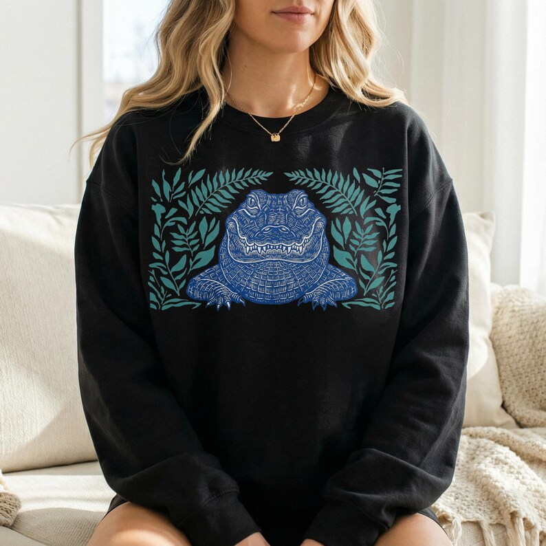 K&ouml;nnte beinhalten: Schwarzes Sweatshirt mit einem detaillierten blauen Alligator-Motiv, umgeben von t&uuml;rkisfarbenen Bl&auml;ttern. Der Alligator weist komplizierte Muster auf und ist mittig platziert. Das Design ist eine Grafik zum Thema Natur.