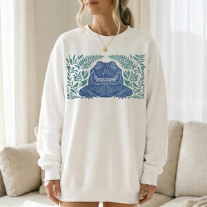 K&ouml;nnte beinhalten: Wei&szlig;es Sweatshirt mit einem blauen Alligator-Design, umrahmt von gr&uuml;nen Bl&auml;ttern. Der Alligator hat detaillierte Muster. Das Sweatshirt ist ein l&auml;ssiger Stil, geeignet f&uuml;r den Alltag.