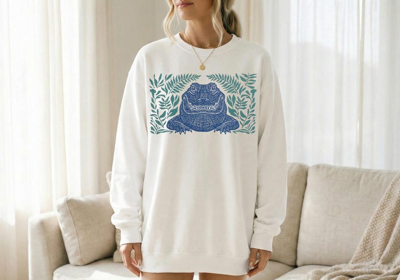 K&ouml;nnte beinhalten: Wei&szlig;es Sweatshirt mit einem blauen Alligator-Design, umrahmt von gr&uuml;nen Bl&auml;ttern. Der Alligator hat detaillierte Muster. Das Sweatshirt ist ein l&auml;ssiger Stil, geeignet f&uuml;r den Alltag.