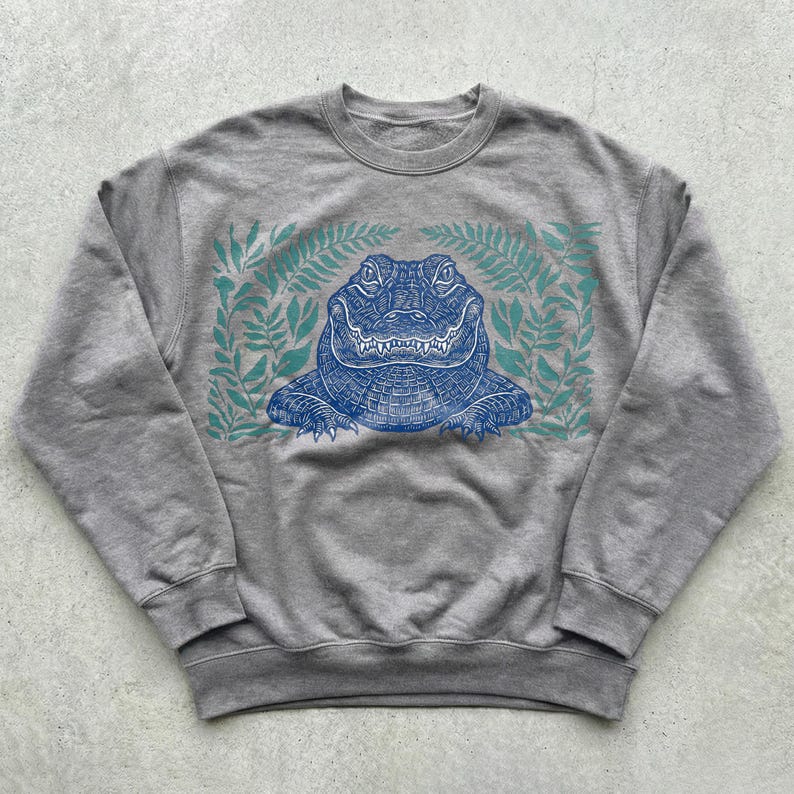K&ouml;nnte beinhalten: Graues Sweatshirt mit einem detaillierten blauen Alligator-Design, umgeben von t&uuml;rkisfarbenen Farnbl&auml;ttern. Der Alligator hat komplizierte Muster und einen l&auml;chelnden Ausdruck, was ein einzigartiges Design ergibt.