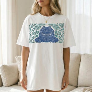 K&ouml;nnte beinhalten: Wei&szlig;es T-Shirt mit einem blauen Alligator-Design, umgeben von gr&uuml;nen Bl&auml;ttern. Der Alligator hat detaillierte Muster. Das Shirt hat eine lockere, &uuml;bergro&szlig;e Passform, geeignet f&uuml;r den Alltag. Das Design ist mittig auf der Vorderseite.
