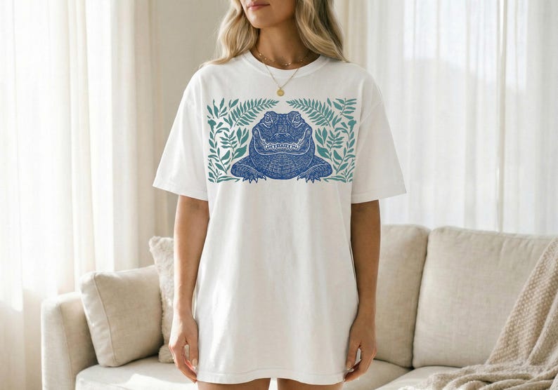 K&ouml;nnte beinhalten: Wei&szlig;es T-Shirt mit einem blauen Alligator-Design, umgeben von gr&uuml;nen Bl&auml;ttern. Der Alligator hat detaillierte Muster. Das Shirt hat eine lockere, &uuml;bergro&szlig;e Passform, geeignet f&uuml;r den Alltag. Das Design ist mittig auf der Vorderseite.