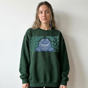 K&ouml;nnte beinhalten: Dunkelgr&uuml;nes Sweatshirt mit einem blauen Alligator-Design und Bl&auml;ttern. Der Alligator ist detailliert, und die Bl&auml;tter umrahmen das Design. Das Sweatshirt hat eine einfarbige Farbe, und das Design ist auf der Vorderseite zentriert.