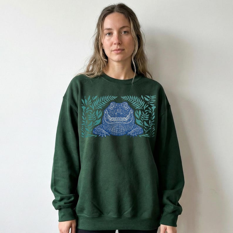 K&ouml;nnte beinhalten: Dunkelgr&uuml;nes Sweatshirt mit einem blauen Alligator-Design und Bl&auml;ttern. Der Alligator ist detailliert, und die Bl&auml;tter umrahmen das Design. Das Sweatshirt hat eine einfarbige Farbe, und das Design ist auf der Vorderseite zentriert.
