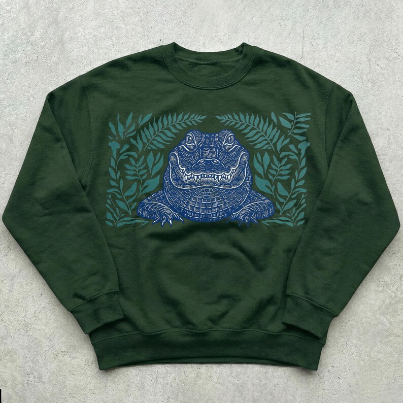 K&ouml;nnte beinhalten: Dunkelgr&uuml;nes Sweatshirt mit einem blauen Alligator-Design, umgeben von gr&uuml;nen Bl&auml;ttern. Der Alligator ist detailliert mit einem strukturierten Muster. Das Sweatshirt mit Rundhalsausschnitt ist f&uuml;r den l&auml;ssigen Gebrauch geeignet.
