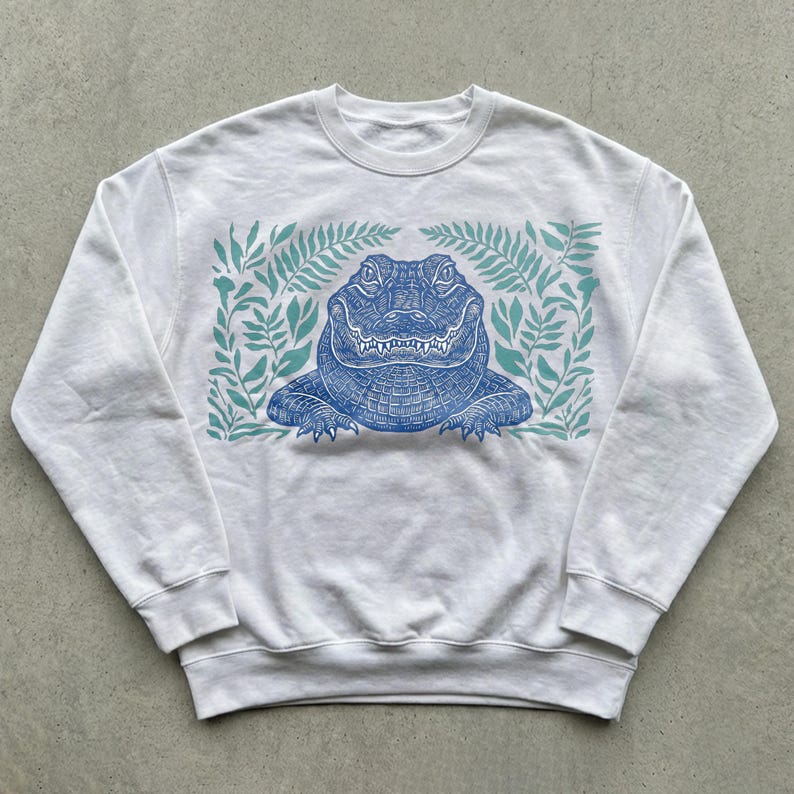 K&ouml;nnte beinhalten: Wei&szlig;es Sweatshirt mit Rundhalsausschnitt und blauem Alligatormotiv, umgeben von t&uuml;rkisfarbenen Bl&auml;ttern. Der Alligator ist detailliert dargestellt. Ein l&auml;ssiges Unisex-Oberteil f&uuml;r den Alltag.