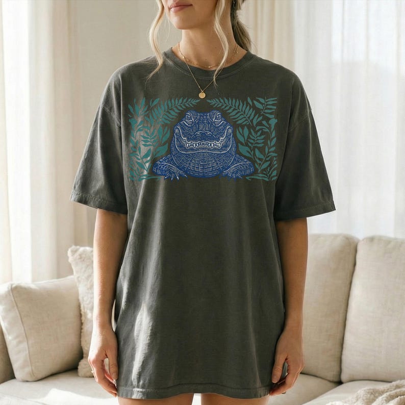 K&ouml;nnte beinhalten: Dunkelgraues T-Shirt mit einem blauen Alligator-Design, umgeben von t&uuml;rkisfarbenen Bl&auml;ttern. Der Alligator ist mit feinen Linien detailliert, und das Shirt hat eine lockere Passform. Das Design ist mittig auf der Vorderseite.