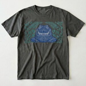 K&ouml;nnte beinhalten: Dunkelgraues T-Shirt mit einer blauen Alligator-Illustration, umgeben von teal-farbenen farnartigen Bl&auml;ttern. Der Alligator hat detaillierte Schuppen und ein grinsendes Maul. Das Shirt ist ein klassischer Rundhals-Stil.