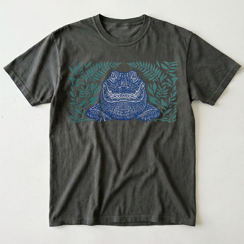 K&ouml;nnte beinhalten: Dunkelgraues T-Shirt mit einer blauen Alligator-Illustration, umgeben von teal-farbenen farnartigen Bl&auml;ttern. Der Alligator hat detaillierte Schuppen und ein grinsendes Maul. Das Shirt ist ein klassischer Rundhals-Stil.